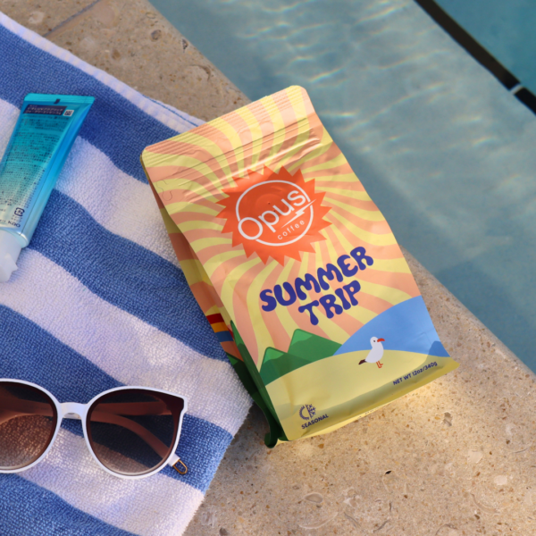 Opus Summer Trip pictured on a beach towel with sunglasses and next to to a pool.