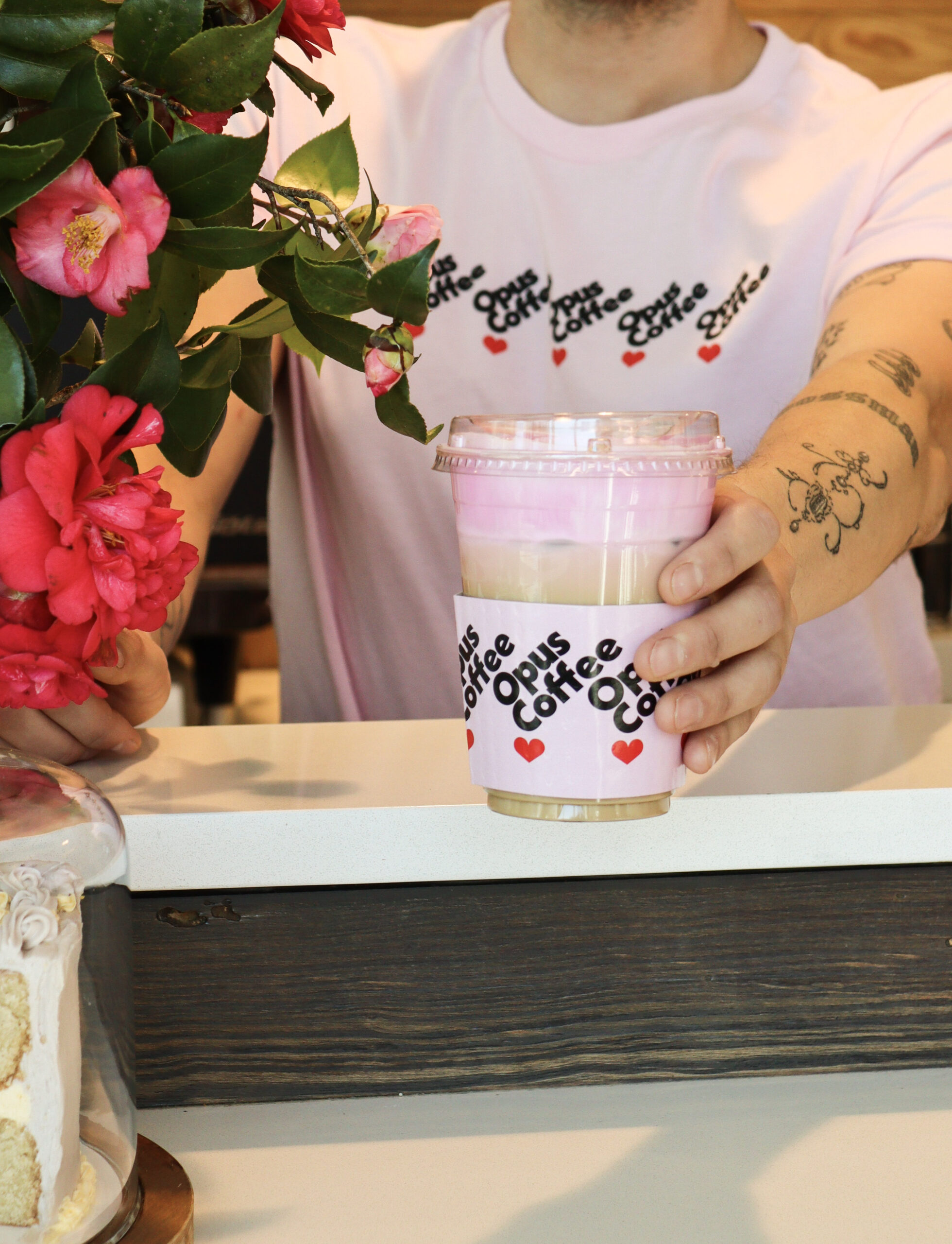 barista wearing pink heart drip shirt handing out drink with flowers in the frame.