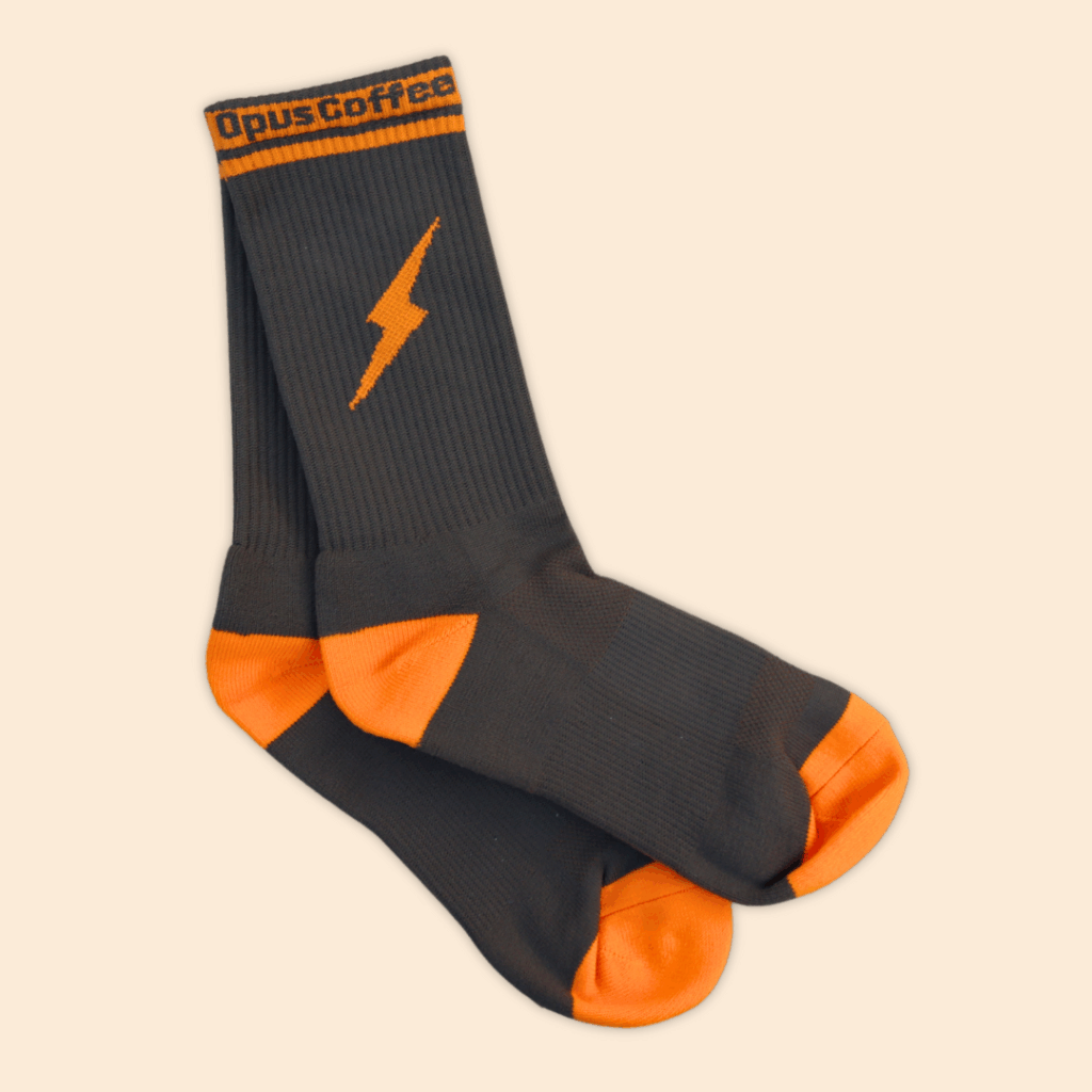 Brown Socks with an orange toe and heel on the foot. There is an orange band around the top that says Opus Coffee in brown and an orange lightning bolt on the side.