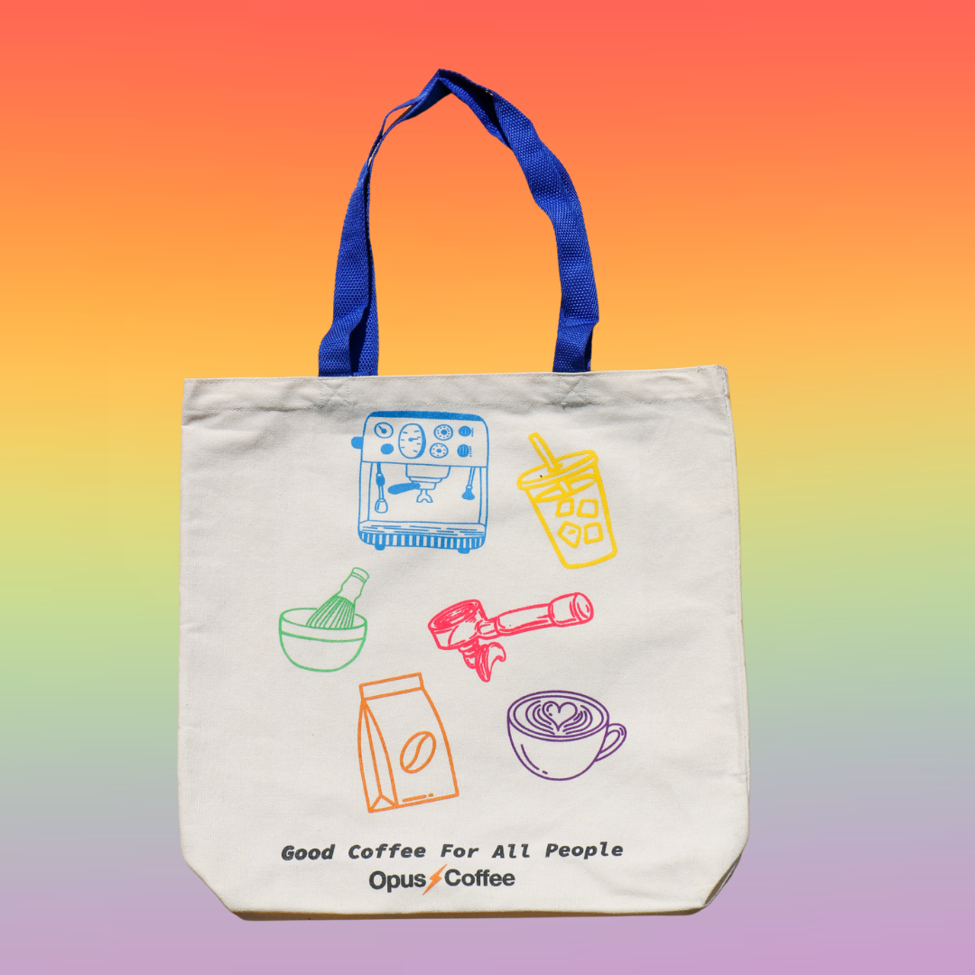 Opus pride tote on rainbow background. Tote is a natural color with blue handles that reads, "good coffee for all people" and has the opus logo. Also pictured on the tote are rainbow colored espresso machine, matcha bowl and whisk, portafilter, cup of coffee with latte art, bag of coffee beans, and iced coffee.